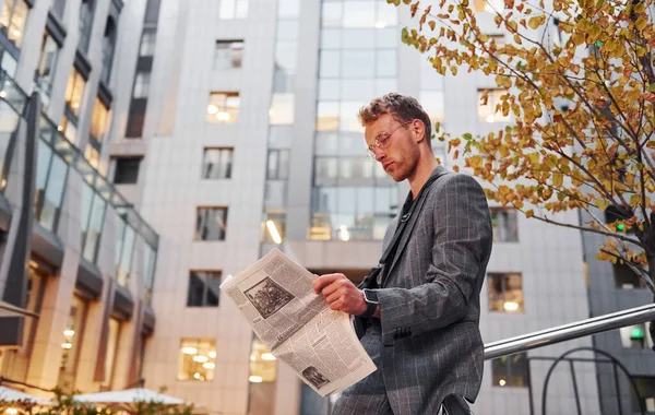 Reads newspaper. Young elegant man in good clothes is outdoors in the city at daytime.