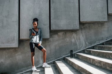 Telefon kullanıyor. Spor elbiseli genç Afrikalı Amerikalı kadın gündüzleri dışarıda antrenman yapıyor..