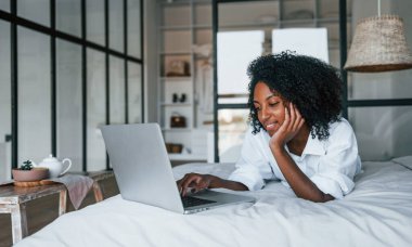 Dizüstü bilgisayarla. Evde kıvırcık saçlı, Afro-Amerikalı bir kadın var..