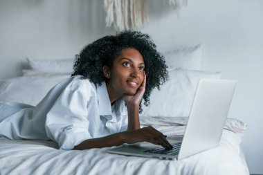 Dizüstü bilgisayarla. Evde kıvırcık saçlı, Afro-Amerikalı bir kadın var..