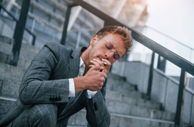 Sigara içiyorum. Gri üniformalı genç işadamı şehirde açık havada..