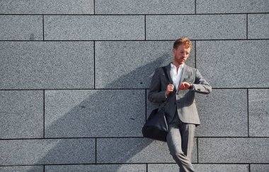 Gri duvara yaslan. Gri üniformalı genç işadamı şehirde açık havada..