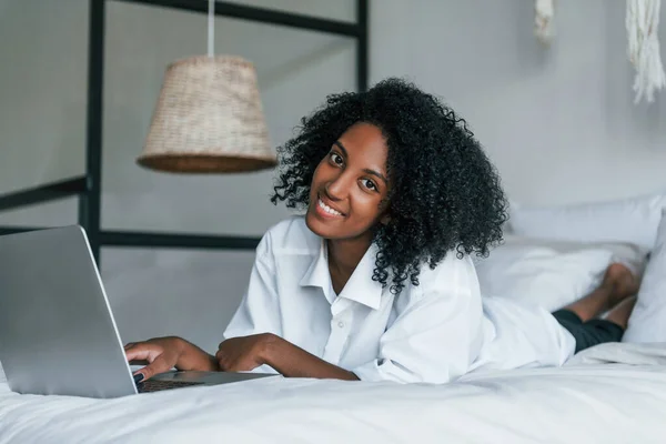 Lying down on bed with laptop. Young african american woman with curly hair indoors at home.