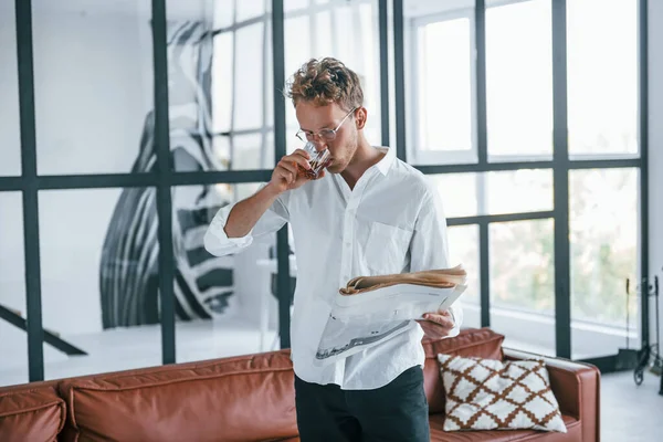 With drink and newspaper. Caucasian young guy in elegant white shirt indoors at home.