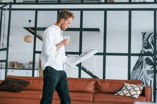 With drink and newspaper. Caucasian young guy in elegant white shirt indoors at home.