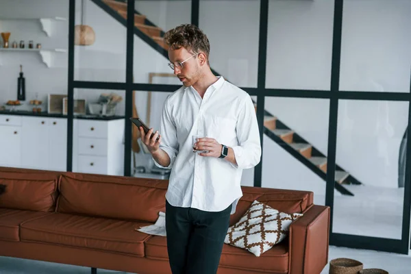 Holds phone and glass. Caucasian young guy in elegant white shirt indoors at home.