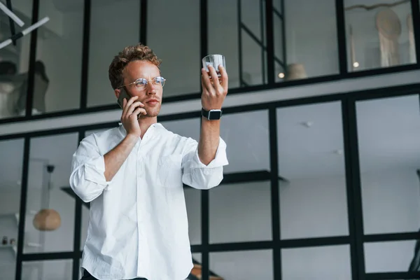 Holds phone and glass. Caucasian young guy in elegant white shirt indoors at home.