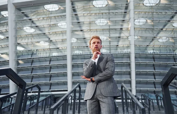 On the stairs of stadium. Young businessman in grey formal wear is outdoors in the city.