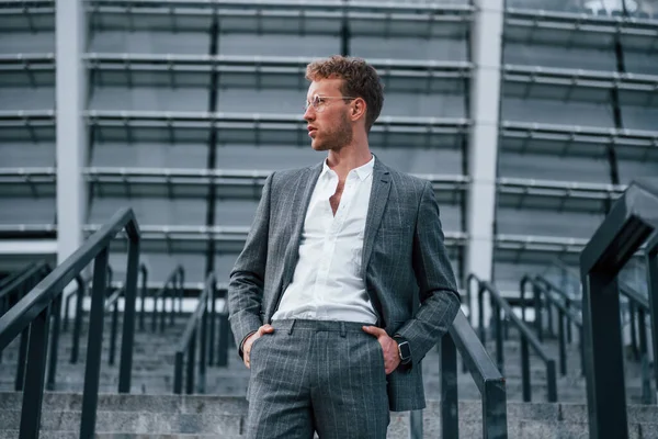 On the stairs of stadium. Young businessman in grey formal wear is outdoors in the city.