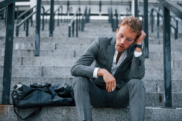 Smoking cigarette. Young businessman in grey formal wear is outdoors in the city.