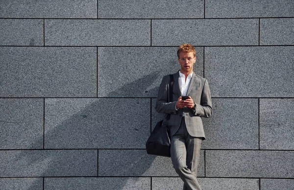 Against grey wall. Young businessman in grey formal wear is outdoors in the city.