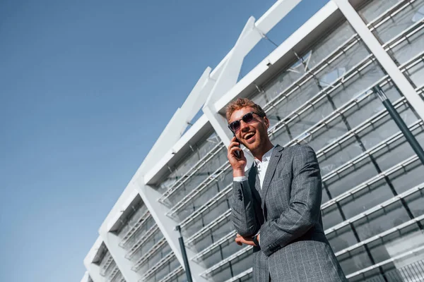 Big stadium behind. Young businessman in grey formal wear is outdoors in the city.