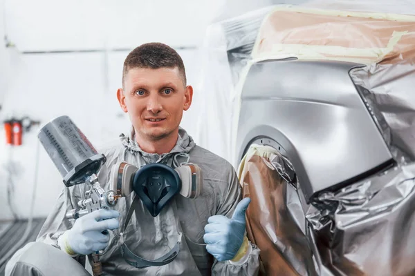 Sits near freshly painted car. Caucasian automobile repairman in uniform works in garage.