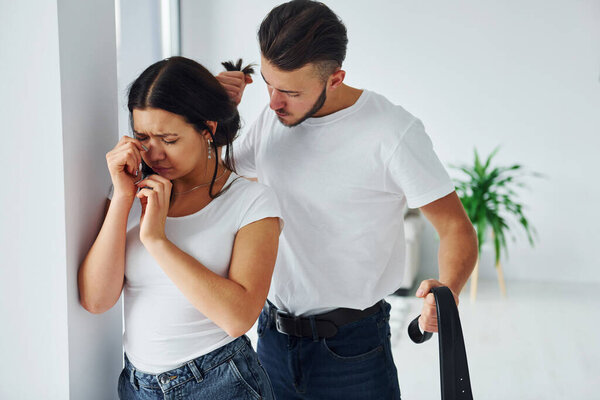 Жестокий агрессивный молодой человек с поясом угрожает своей девушке дома.