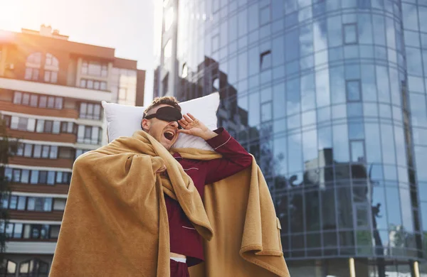 Against business building. Young man in pajama is outdoors on the street. Feels sleepy.