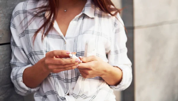 Positive young woman in casual clothes standing outdoors near wall with modern digital cigarette.