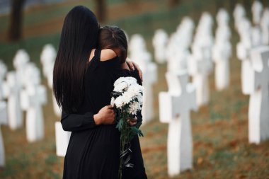 Sarılmak ve ağlamak. Siyah elbiseli iki genç kadın bir sürü beyaz haçla mezarlığı ziyaret ediyor. Cenaze ve ölüm kavramı.