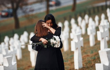 Sarılmak ve ağlamak. Siyah elbiseli iki genç kadın bir sürü beyaz haçla mezarlığı ziyaret ediyor. Cenaze ve ölüm kavramı.