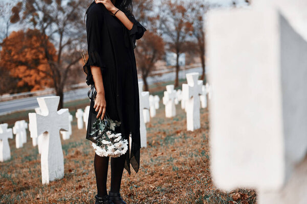 With flowers in hands. Young woman in black clothes visiting cemetery with many white crosses. Conception of funeral and death.