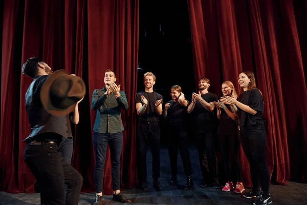 Standing against red curtains. Group of actors in dark colored clothes on rehearsal in the theater.