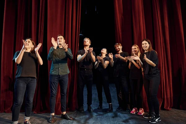 Standing against red curtains. Group of actors in dark colored clothes on rehearsal in the theater.