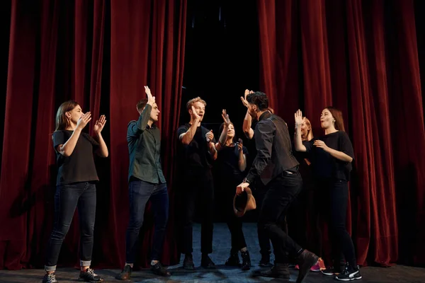 Standing against red curtains. Group of actors in dark colored clothes on rehearsal in the theater.