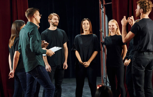 Стоя напротив красных занавесок. Группа актёров в тёмной одежде на репетиции в театре.