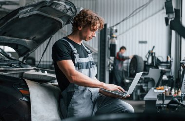 Dizüstü bilgisayarla. Gri üniformalı yetişkin adam otomobil salonunda çalışıyor..