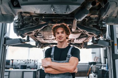 Gri üniformalı yakışıklı yetişkin adam otomobilin altında duruyor..
