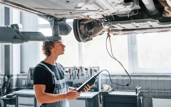 Special tool for testing characteristics. Adult man in grey colored uniform works in the automobile salon.