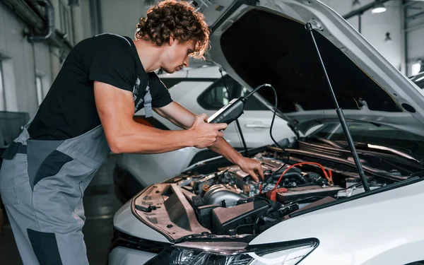 Tests car's electronics. Adult man in grey colored uniform works in the automobile salon.