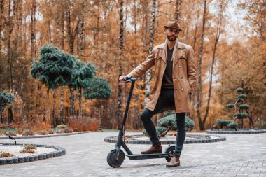 Parkta, elektrikli scooter 'la. Şık giyimli genç erkek model gündüz vakti dışarıda parkta..