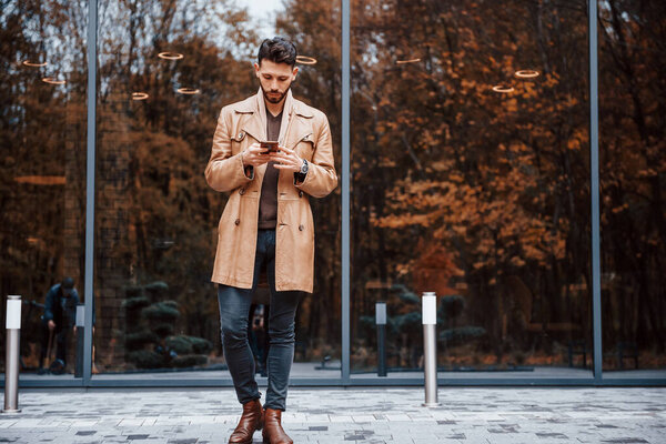 Holds smartphone. Young male model in fashionable clothes is outdoors in the city at daytime.