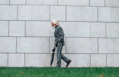 Gri saçlı ve sakallı kıdemli işadamı dışarıda elinde şemsiyeyle yürüyor..