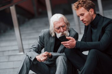 Telefonla oturuyor. Zarif giyinmiş son sınıf öğrencisi olan genç adam açık havada birlikte. İş kavramı.