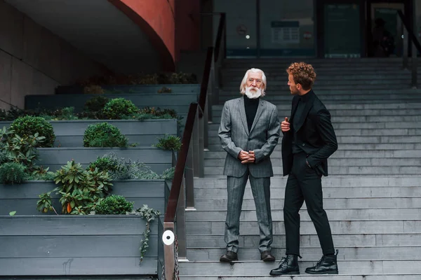 Walking on the stairs. Young guy with senior man in elegant clothes is outdoors together. Conception of business.