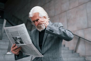 Gazete okuyor. Gri saçlı ve sakallı kıdemli işadamı dışarıda..