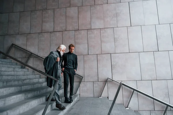 Walking on the stairs. Young guy with senior man in elegant clothes is outdoors together. Conception of business.