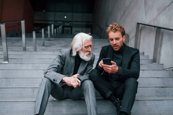 Sitting with phone. Young guy with senior man in elegant clothes is outdoors together. Conception of business.