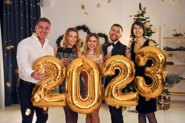2023 numaralı balonları tutuyorum. Bir grup insan içeride yeni yıl partisi veriyor..