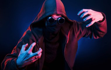 Gözlüklü, maskeli ve başlıklı korkunç adam. Neon ışıkları. Genç Avrupalı adam karanlık stüdyoda..