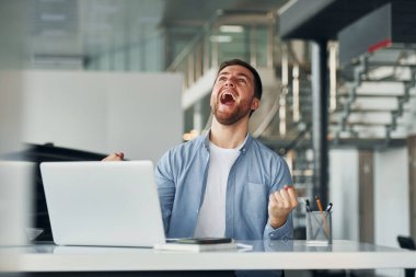 Laptop kullanıyor. Beyaz gömlekli ve mavi ceketli genç adam içeride çalışıyor..