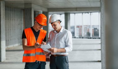 Takım elbiseli bir adam ve turuncu giysili bir tamirci inşaatta çalışıyor..