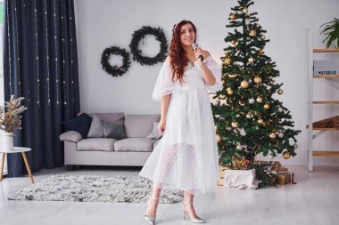 Beyaz elbiseli ve mikrofonlu bir kadın Noel süslemeli yurt odasında şarkı söylüyor..