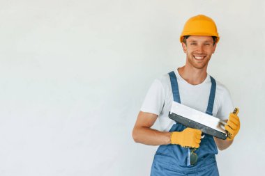 Beyaz duvarın yanında duruyor. Gündüz vakti inşaatta üniformalı genç bir adam çalışıyor..