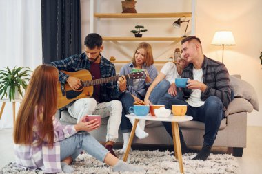 Akustik gitar çalıyor. Bir grup arkadaş içeride parti veriyor..