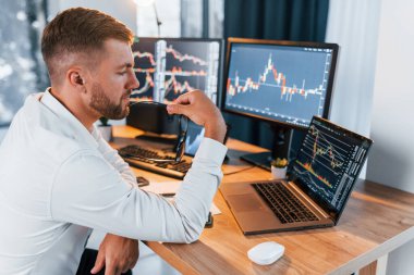 Bilgi analiz ediliyor. Resmi giyinmiş genç işadamı birden fazla ekranla ofiste..