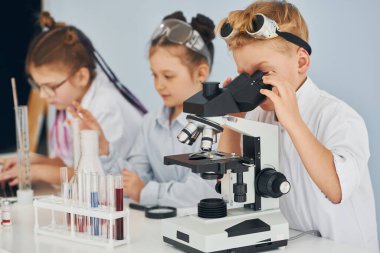 Mikroskop kullanarak. Beyaz önlüklü çocuklar laboratuarda ekipman kullanarak bilim adamlarını oynuyorlar..