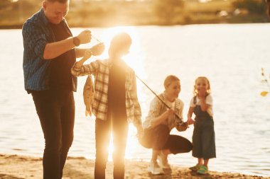Güneş ışığıyla aydınlatılmış. Baba ve anne oğul ve kız yazın birlikte açık havada balık tutuyorlar..
