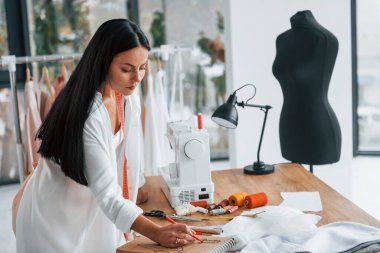 Masanın yanında duruyor. Terzi odasında, farklı kıyafetlerle..